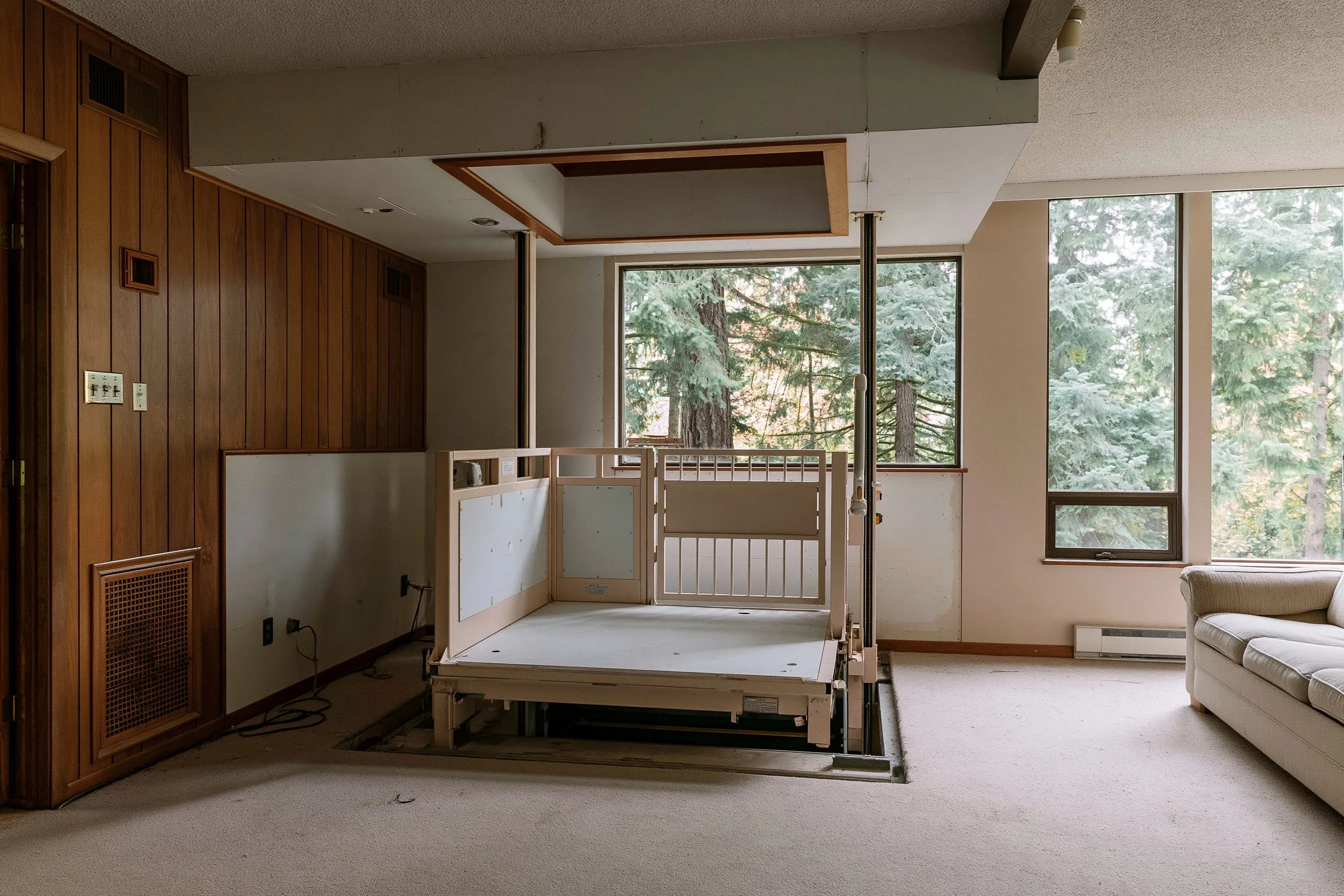 Wide platform lift with low side panels and a gate in the corner of a wood-paneled family room, under a framed opening in the ceiling